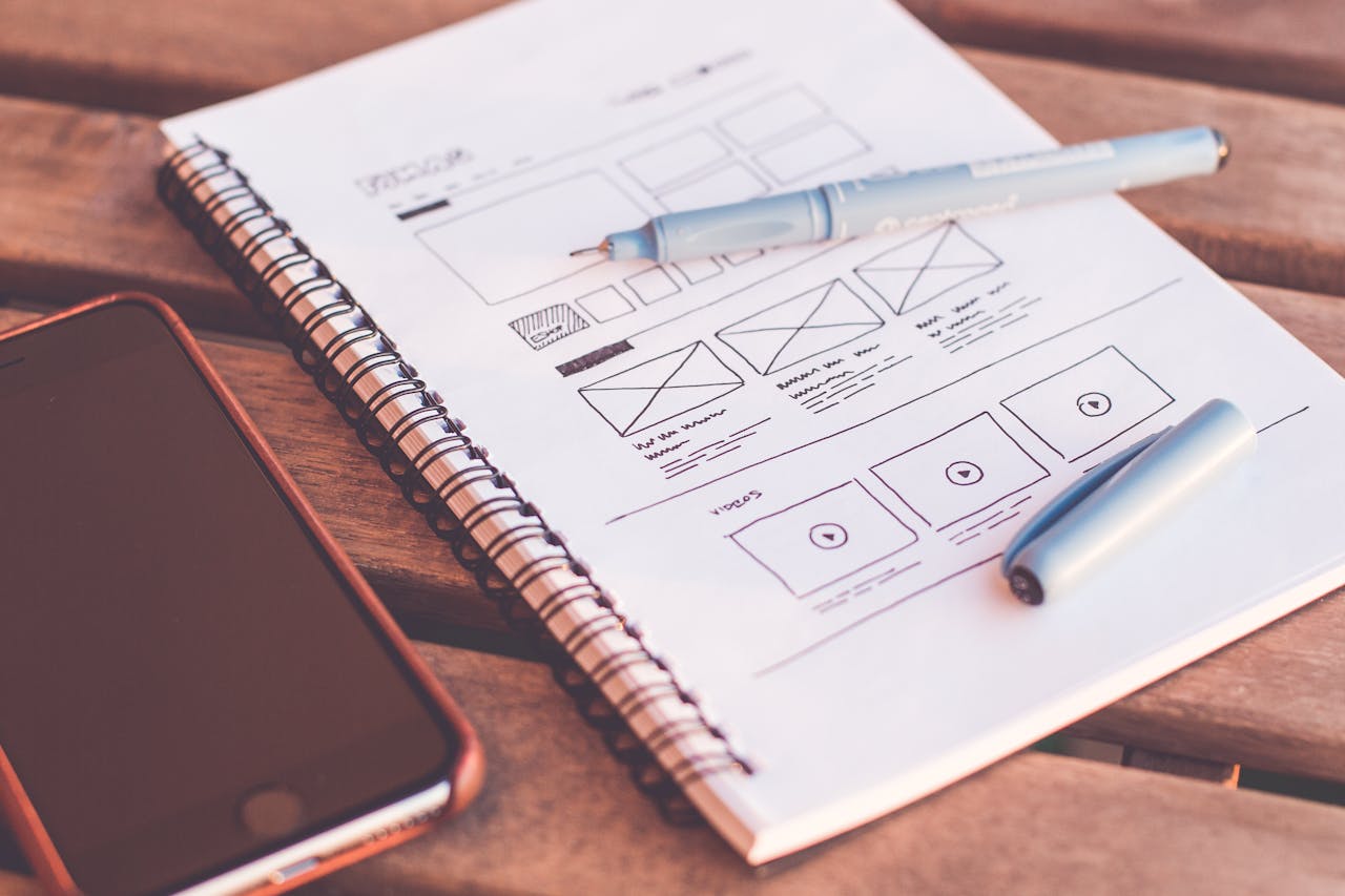 our-journey-01 Close-up of a notebook with wireframe sketches and a smartphone on a wooden desk.